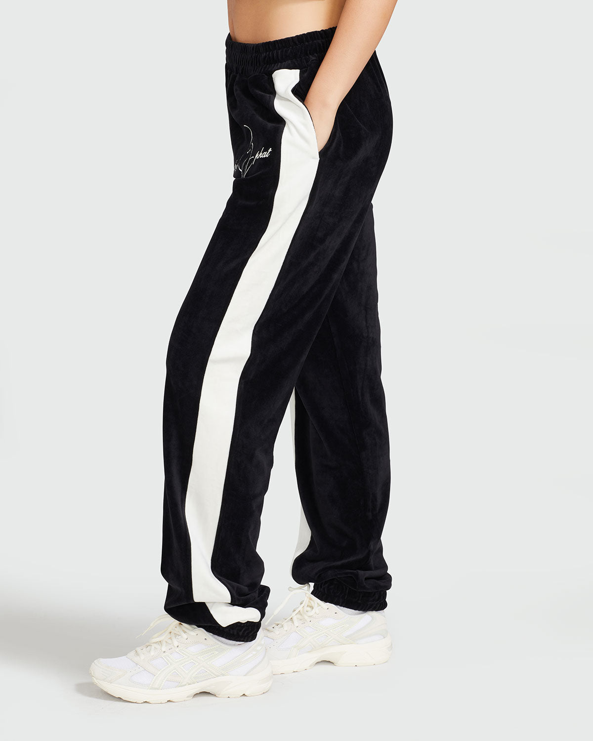 Side view image of standard sized model wearing black velour sweatpant with color blocked cream stripe down both sides. Sweatpant features embroidered Baby Phat script and cat logo on wearer's upper left thigh. Model has her hand in her pocket.