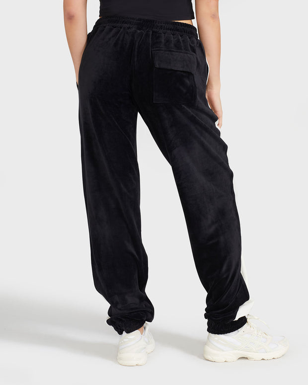 Back view image of standard sized model wearing black velour sweatpant with color blocked cream stripe down both sides. Sweatpant features oversized flap pocket on rear right side.