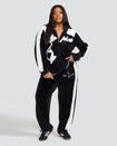 Full body image of plus sized model wearing black velour zip up hoodie with color blocked off white panel down both sleeves. Zippered sweatshirt features off white Baby Phat script logo across upper chest and oversized off white solid cat logo on wearer's right front. Model is also wearing matching black sweatpant with off white color blocking and off white embroidered script and cat logo on wearer's upper left thigh.