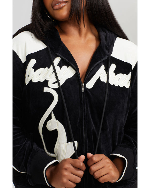 Detail image of plus sized model wearing black velour zip up hoodie with color blocked off white panel down both sleeves. Zippered sweatshirt features off white Baby Phat script logo across upper chest and oversized solid cat logo on wearer's right front. Zippered sweatshirt also features matching black ribbing at the wrists and hem with off white tipping.