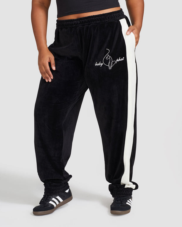 Plus sized model shown wearing black velour sweatpant with color blocked cream stripe down both sides. Sweatpant features embroidered Baby Phat script and cat logo on wearer's upper left thigh. Model has hand in her pocket.