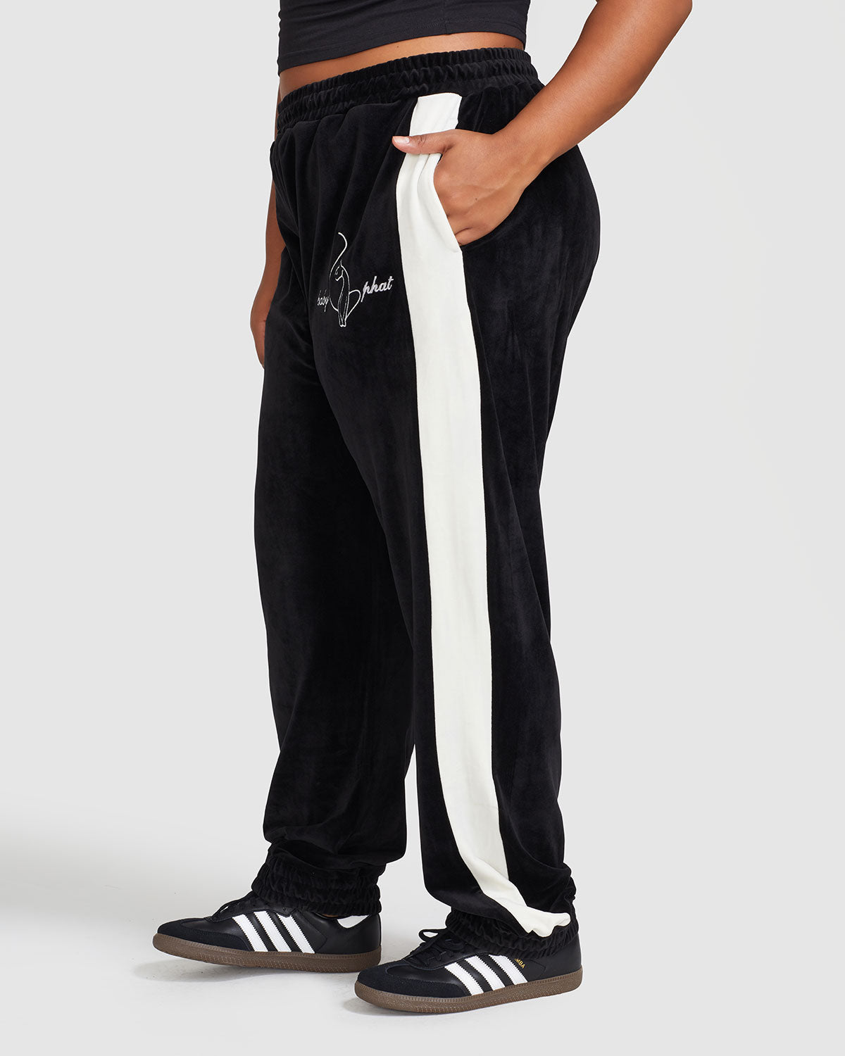 Side view image of plus sized model wearing black velour sweatpant with color blocked cream stripe down both sides. Sweatpant features embroidered Baby Phat script and cat logo on wearer's upper left thigh. Model has her hand in her pocket.