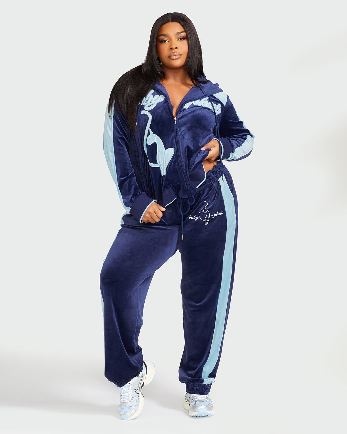 Full body image of plus sized model wearing navy blue velour zip up hoodie with color blocked light blue panel down both sleeves. Zippered sweatshirt features light blue Baby Phat script logo across upper chest and oversized light blue solid cat logo on wearer's right front. Model is also wearing matching navy blue sweatpant with light color blocking and light blue embroidered script and cat logo on wearer's upper left thigh.