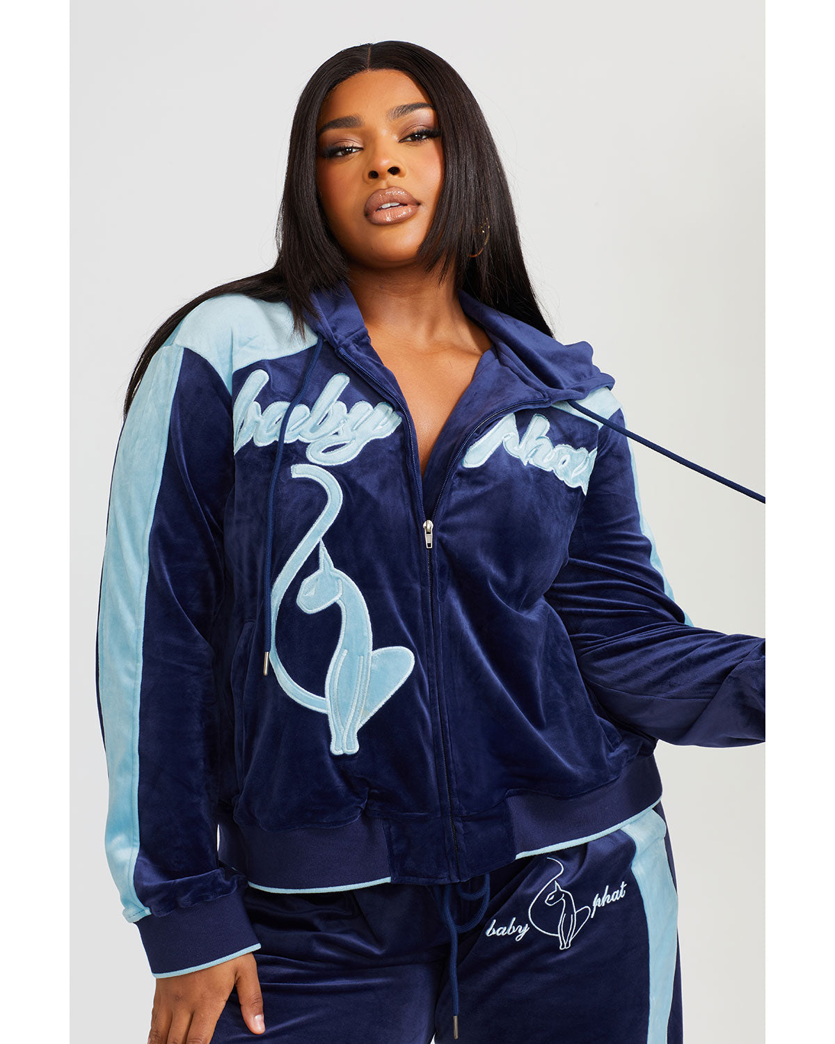 Plus sized model shown wearing navy blue velour zip up hoodie with color blocked light blue panel down both sleeves. Zippered sweatshirt features light blue Baby Phat script logo across upper chest and oversized solid cat logo on wearer's right front. Model is also wearing matching navy blue sweatpant with light blue color blocking. 