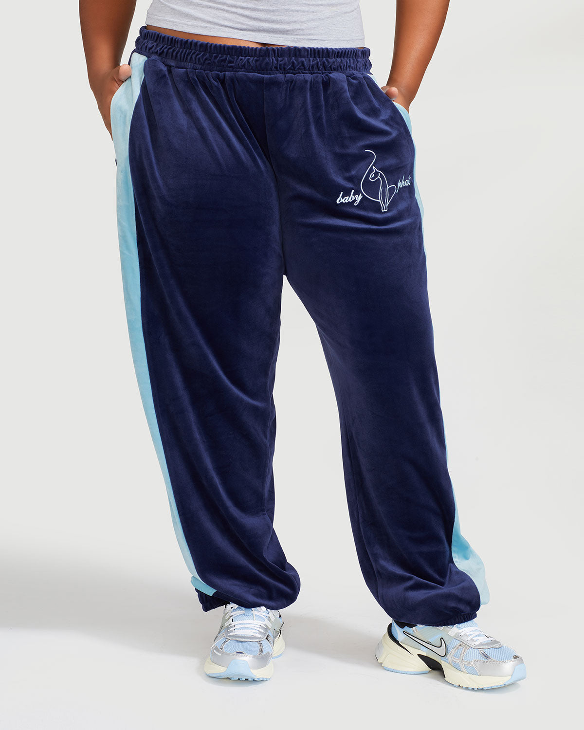 Plus sized model shown wearing navy blue velour sweatpant with color blocked light blue stripe down both sides. Sweatpant features embroidered Baby Phat script and cat logo on wearer's upper left thigh. Model has both hands in her pockets.