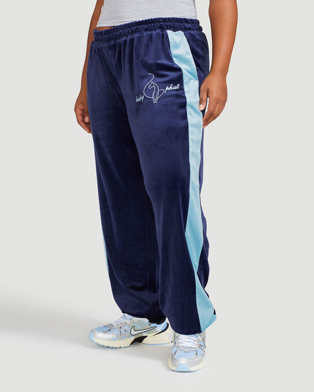 Side view image shows plus sized model wearing navy blue velour sweatpant with color blocked light blue stripe down both sides. Sweatpant features embroidered Baby Phat script and cat logo on wearer's upper left thigh. 