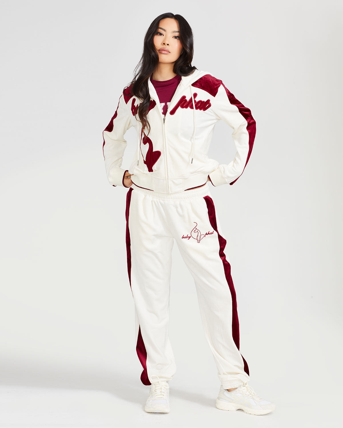 Full body image of standard sized model wearing off white velour zip up hoodie with color blocked burgundy panel down both sleeves. Zippered sweatshirt features burgundy Baby Phat script logo across upper chest and oversized burgundy solid cat logo on wearer's right front. Model is also wearing matching off white sweatpant with burgundy color blocking and burgundy embroidered script and cat logo on wearer's upper left thigh.