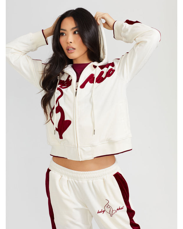 Standard sized model shown wearing off white velour zip up hoodie with color blocked burgundy panel down both sleeves. Zippered sweatshirt features burgundy Baby Phat script logo across upper chest and oversized solid cat logo on wearer's right front. Model is also wearing matching off white sweatpant with burgundy color blocking and burgundy embroidered script and cat logo on wearer's upper left thigh.