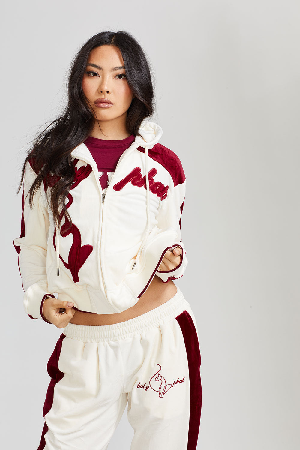 Standard sized model shown wearing off white velour zip up hoodie with color blocked burgundy panel down both sleeves. Zippered sweatshirt features burgundy Baby Phat script logo across upper chest and oversized solid cat logo on wearer's right front. Model is also wearing matching off white sweatpant with burgundy color blocking and burgundy embroidered script and cat logo on wearer's upper left thigh.