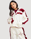 Standard sized model shown wearing off white velour zip up hoodie with color blocked burgundy panel down both sleeves. Zippered sweatshirt features burgundy Baby Phat script logo across upper chest and oversized solid cat logo on wearer's right front. Model is also wearing matching off white sweatpant with burgundy color blocking and burgundy embroidered script and cat logo on wearer's upper left thigh.