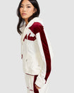 Side view image of standard sized model wearing off white velour zip up hoodie with color blocked burgundy panel down both sleeves. Zippered sweatshirt features matching off white ribbing at the wrists and hem with burgundy tipping. Model is also wearing matching off white sweatpant with burgundy color blocking. 