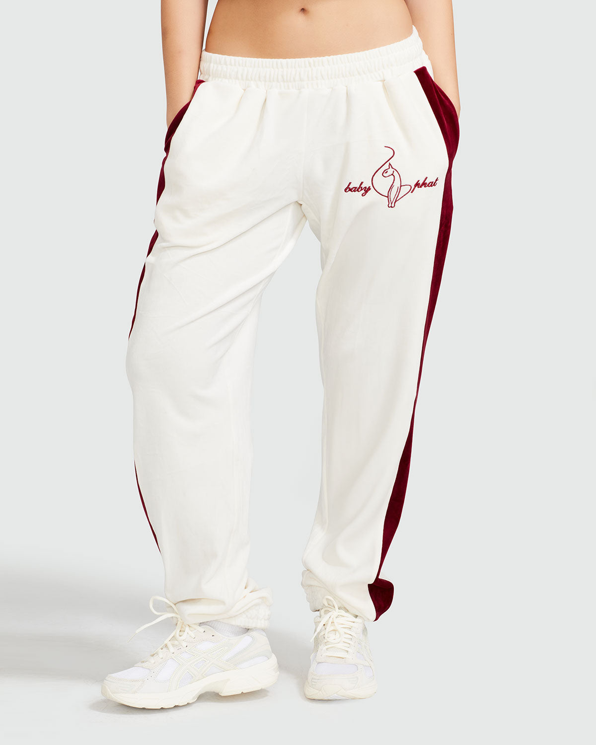 Standard sized model shown wearing off white velour sweatpant with color blocked burgundy stripe down both sides. Sweatpant features embroidered Baby Phat script and cat logo on wearer's upper left thigh. 