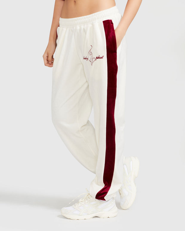 Side view image of standard sized model wearing off white velour sweatpant with color blocked burgundy stripe down both sides. Sweatpant features embroidered Baby Phat script and cat logo on wearer's upper left thigh. Model has hand in her pocket.