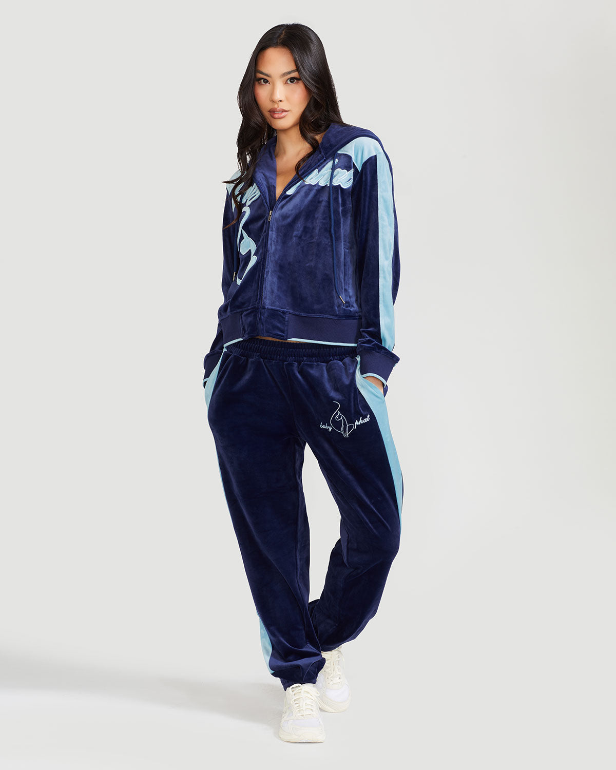 Full body image of standard sized model wearing navy blue velour zip up hoodie with color blocked light blue panel down both sleeves. Zippered sweatshirt features light blue Baby Phat script logo across upper chest and oversized light blue solid cat logo on wearer's right front. Model is also wearing matching navy blue sweatpant with light color blocking and light blue embroidered script and cat logo on wearer's upper left thigh.