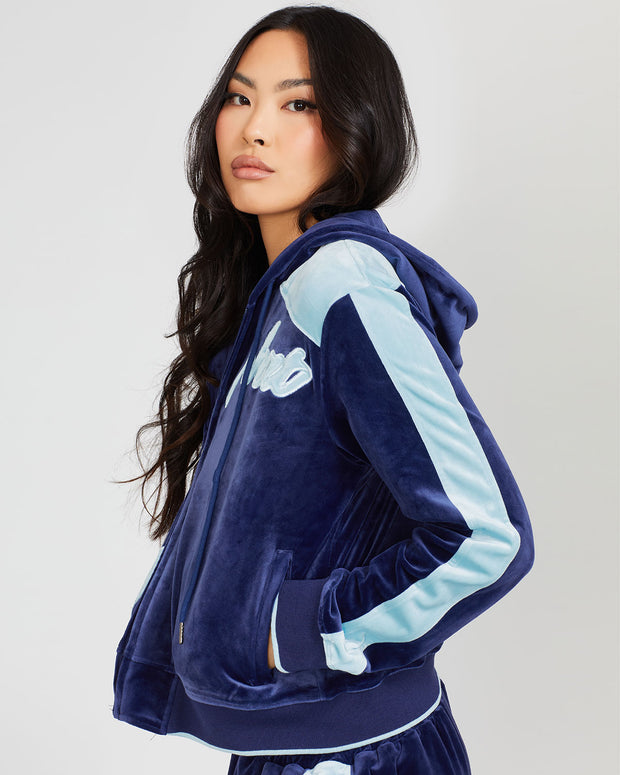 Side view image of standard sized model wearing navy blue velour zip up hoodie with color blocked light blue panel down both sleeves. Zippered sweatshirt features matching navy blue ribbing at the wrists and hem with light blue tipping. Model is also wearing matching navy blue sweatpant with light blue color blocking. 