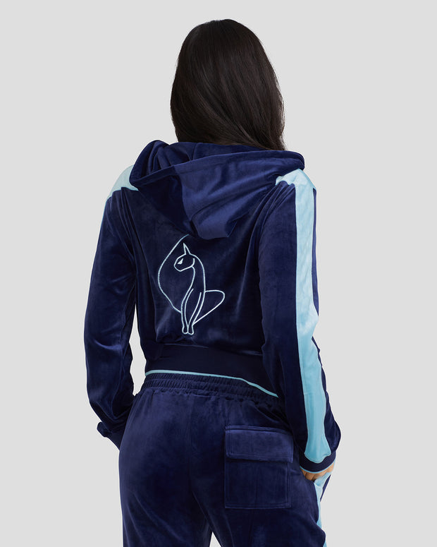 Back view image of standard sized model wearing navy blue velour zip up hoodie with color blocked light blue panel down both sleeves. Zippered sweatshirt features matching navy blue ribbing at the wrists and hem with light blue tipping. Large embroidered cat logo on center back. Model is also wearing matching navy blue sweatpant with light blue color blocking. Hood is down hanging behind wearer's neck. 