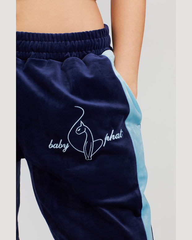 Detail image of standard sized model wearing navy blue velour sweatpant with color blocked light blue stripe down both sides. Sweatpant features embroidered Baby Phat script and cat logo on wearer's upper left thigh. 