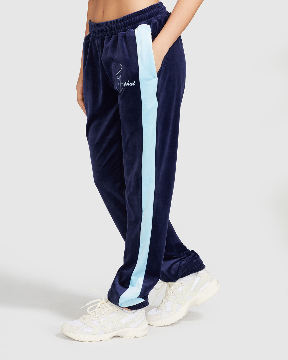 Side view image of standard sized model wearing navy blue velour sweatpant with color blocked light blue stripe down both sides. Sweatpant features embroidered Baby Phat script and cat logo on wearer's upper left thigh. Model has hand in her pocket.