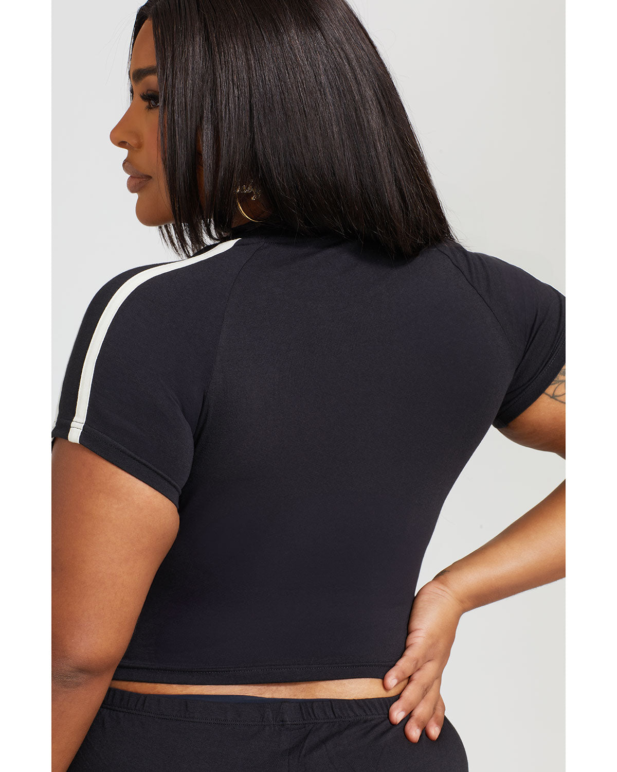 Plus sized model shown wearing black baby tee shirt in this back view image. Shirt features off white stripes on the shoulder. 