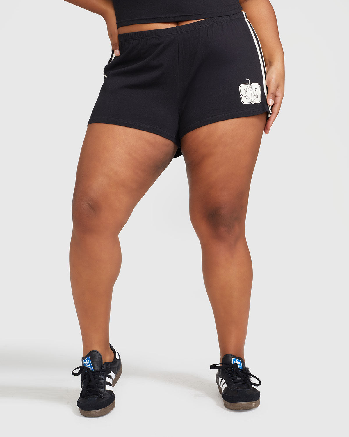 Plus sized model’s legs shown wearing black micro short with 2 contrasting off white stripes down both sides. Short features off white Baby Phat cat 99 heritage logo on wearer's left thigh. 
