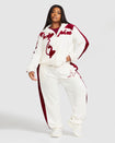 Full body image of plus sized model wearing off white velour zip up hoodie with color blocked burgundy panel down both sleeves. Zippered sweatshirt features burgundy Baby Phat script logo across upper chest and oversized burgundy solid cat logo on wearer's right front. Model is also wearing matching off white sweatpant with burgundy color blocking and burgundy embroidered script and cat logo on wearer's upper left thigh.