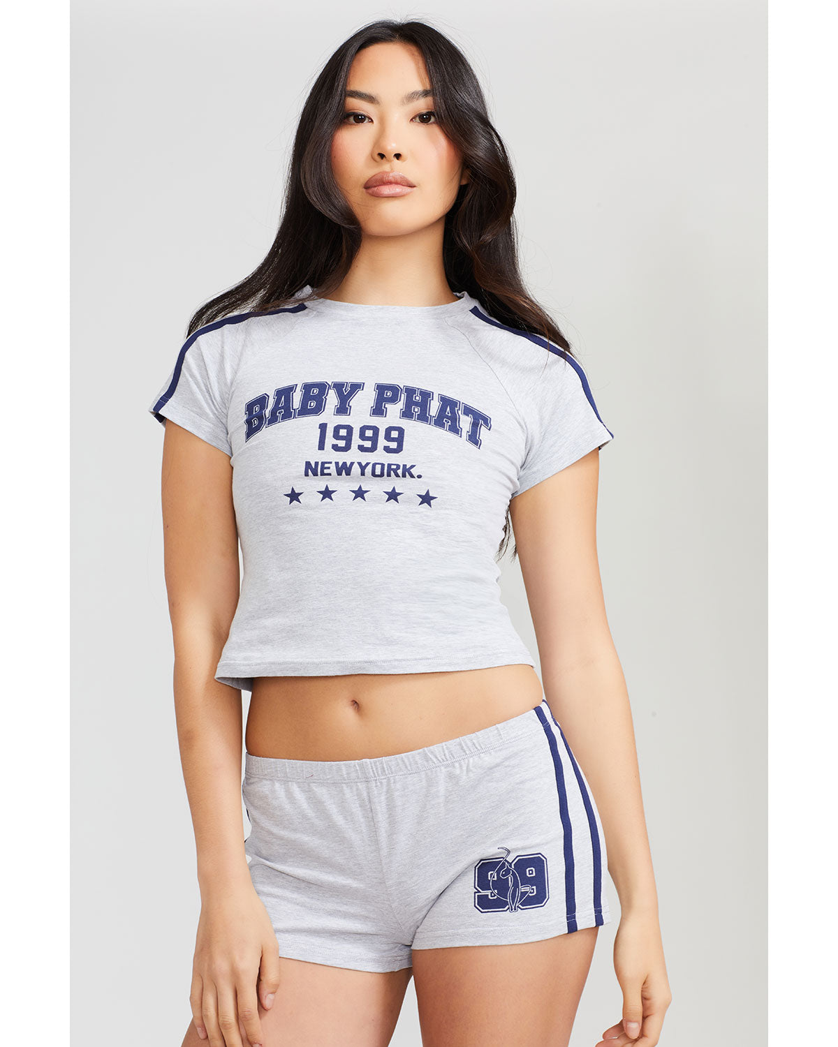 Standard sized model shown wearing heather grey baby tee shirt with navy blue Baby Phat logo, New York, 1999, and stars on the center front. Shirt features navy blue stripes on the shoulder. Model is also wearing matching grey micro shorts with navy blue side stripes and a 99 cat logo.