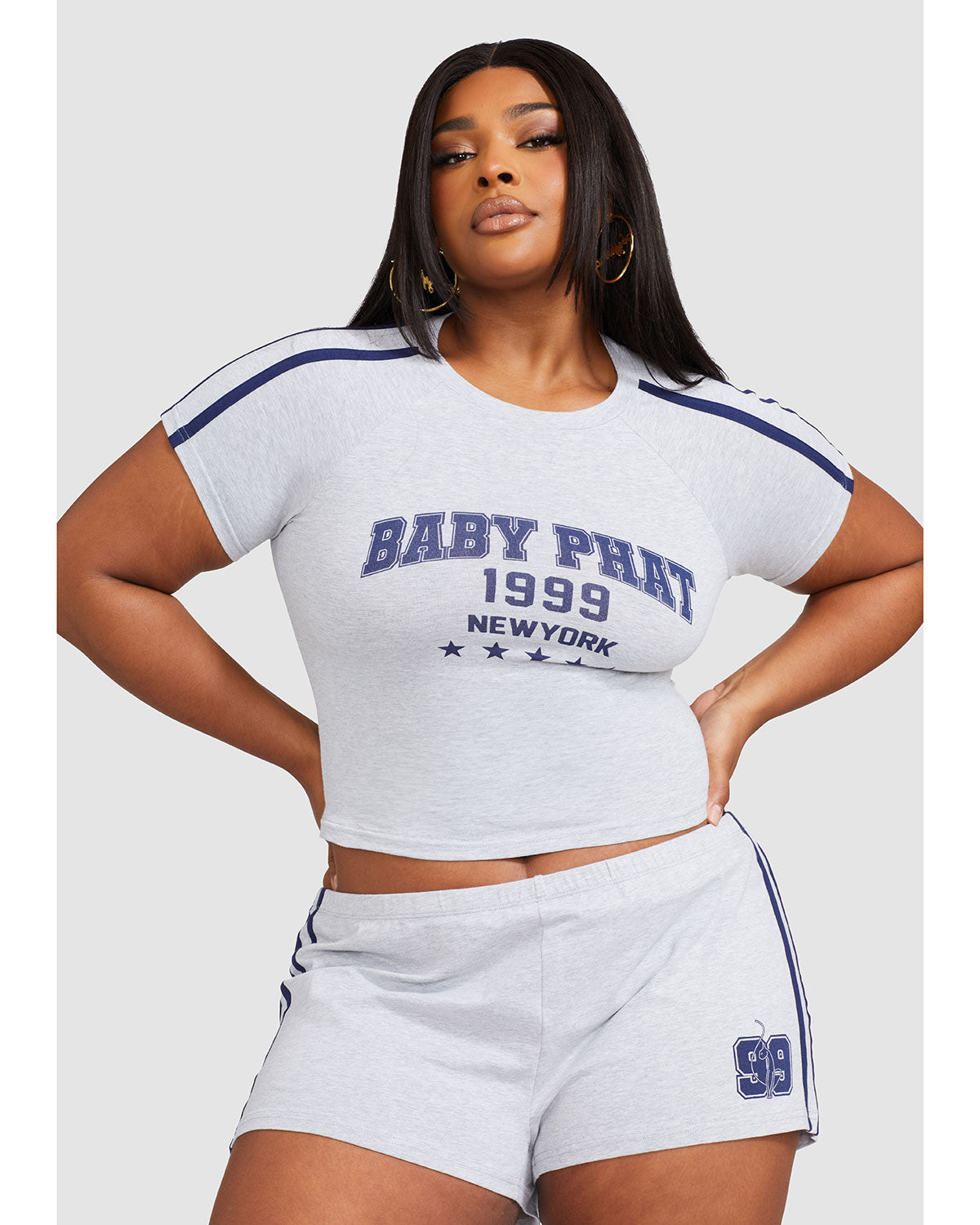 Plus sized model shown wearing heather grey baby tee shirt with navy blue Baby Phat logo, New York, 1999, and stars on the center front. Shirt features navy blue stripes on the shoulder. Model is also wearing matching micro shorts with side stripes and a 99 cat logo.