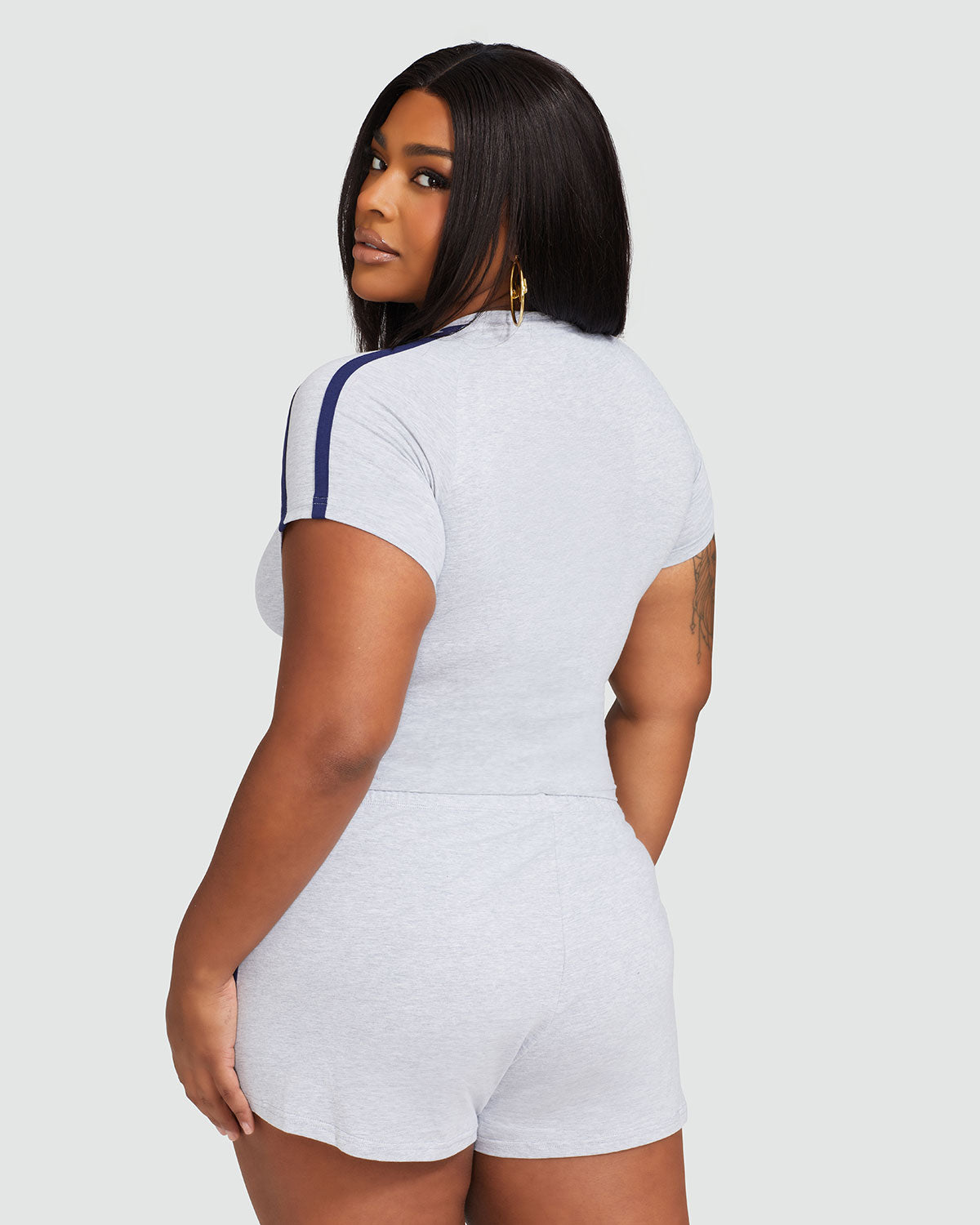 Back view image of plus sized model wearing heather grey baby tee shirt with navy blue stripes on the shoulder. Model is also wearing matching grey micro shorts.