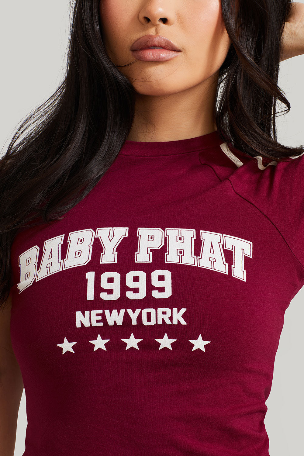 Detail photo shows standard sized model wearing burgundy baby tee shirt with cream Baby Phat logo, New York, 1999, and stars on the center front. 