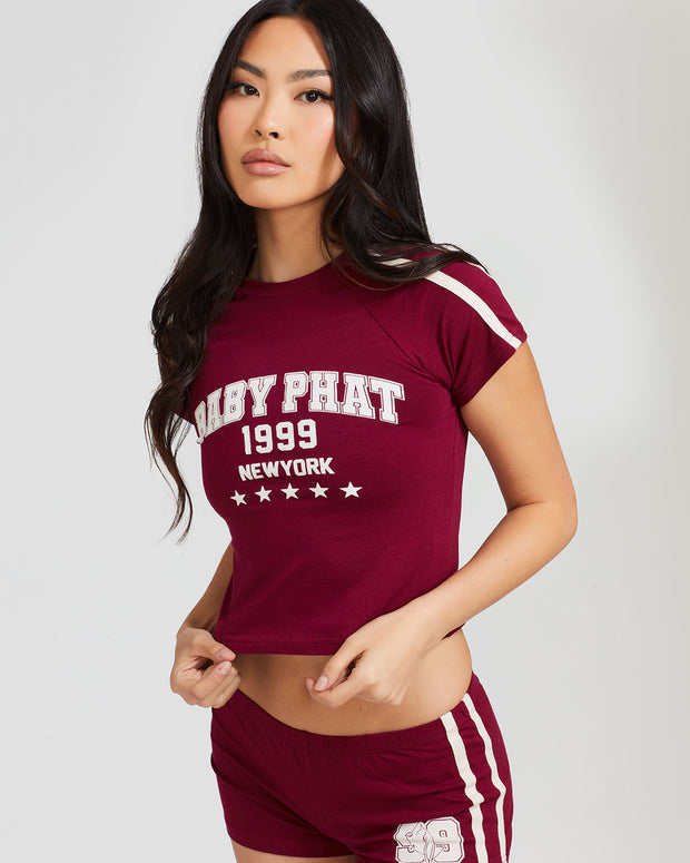 Standard sized model shown wearing burgundy baby tee shirt with cream Baby Phat logo, New York, 1999, and stars on the center front. Shirt features cream stripes on the shoulder. Model is also wearing matching burgundy micro shorts with cream side stripes and a 99 cat logo.