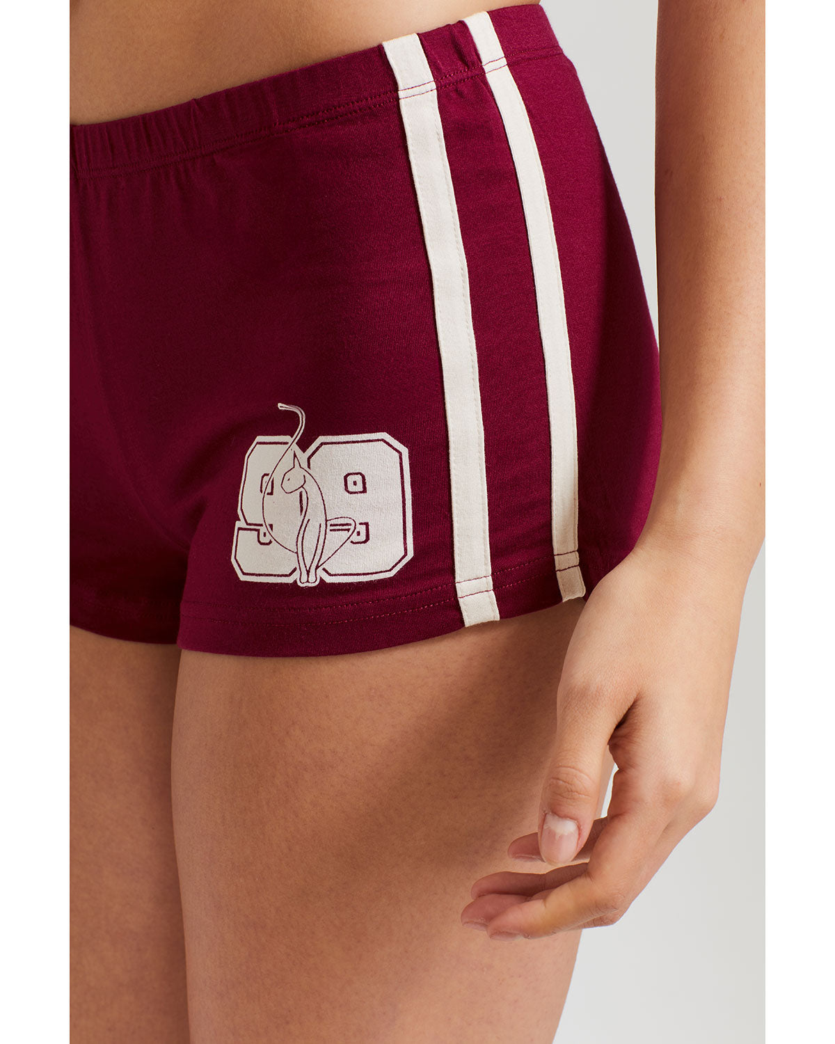 Detail shot of standard sized model’s legs shown wearing burgundy micro short with 2 contrasting off white stripes down both sides. Short features off white Baby Phat cat 99 heritage logo on wearer's left thigh. 
