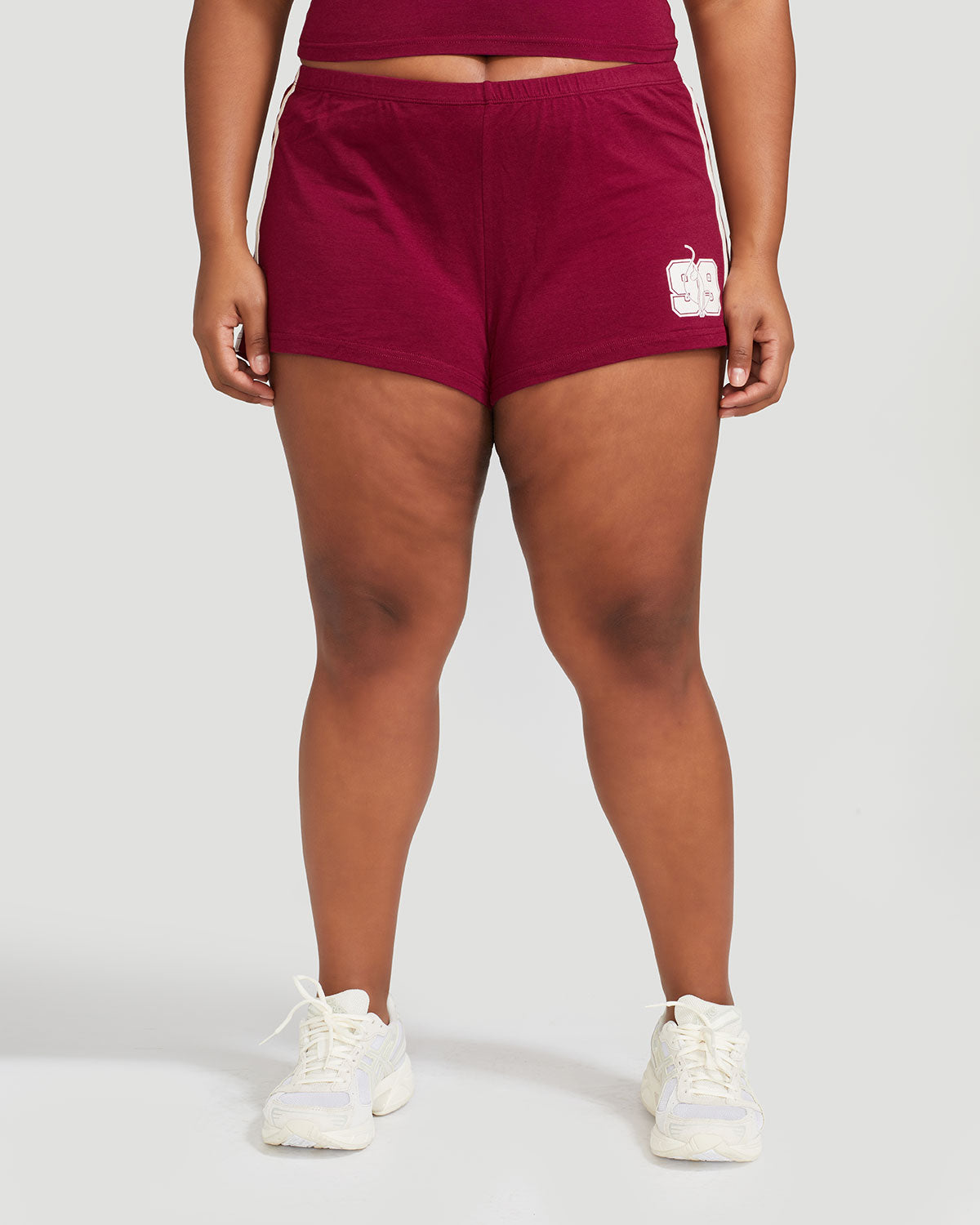 Plus sized model’s legs shown wearing burgundy micro short with 2 contrasting off white stripes down both sides. Short features off white Baby Phat cat 99 heritage logo on wearer's left thigh. 
