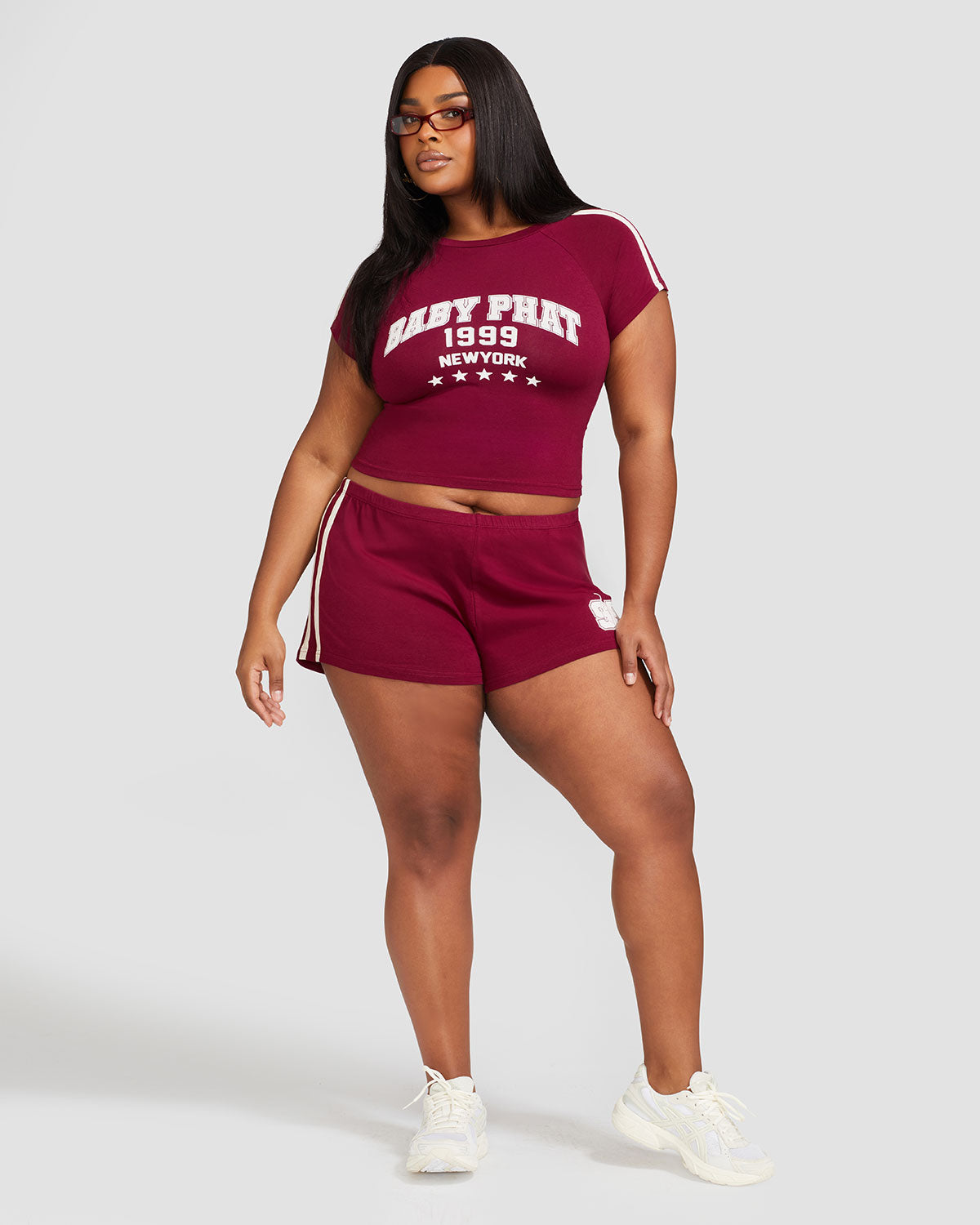 Full body photo shows plus sized model wearing burgundy baby tee shirt with cream Baby Phat logo, New York, 1999, and stars on the center front. Shirt features cream stripes on the shoulder. Model is also wearing matching burgundy micro shorts with cream side stripes and a 99 cat logo.