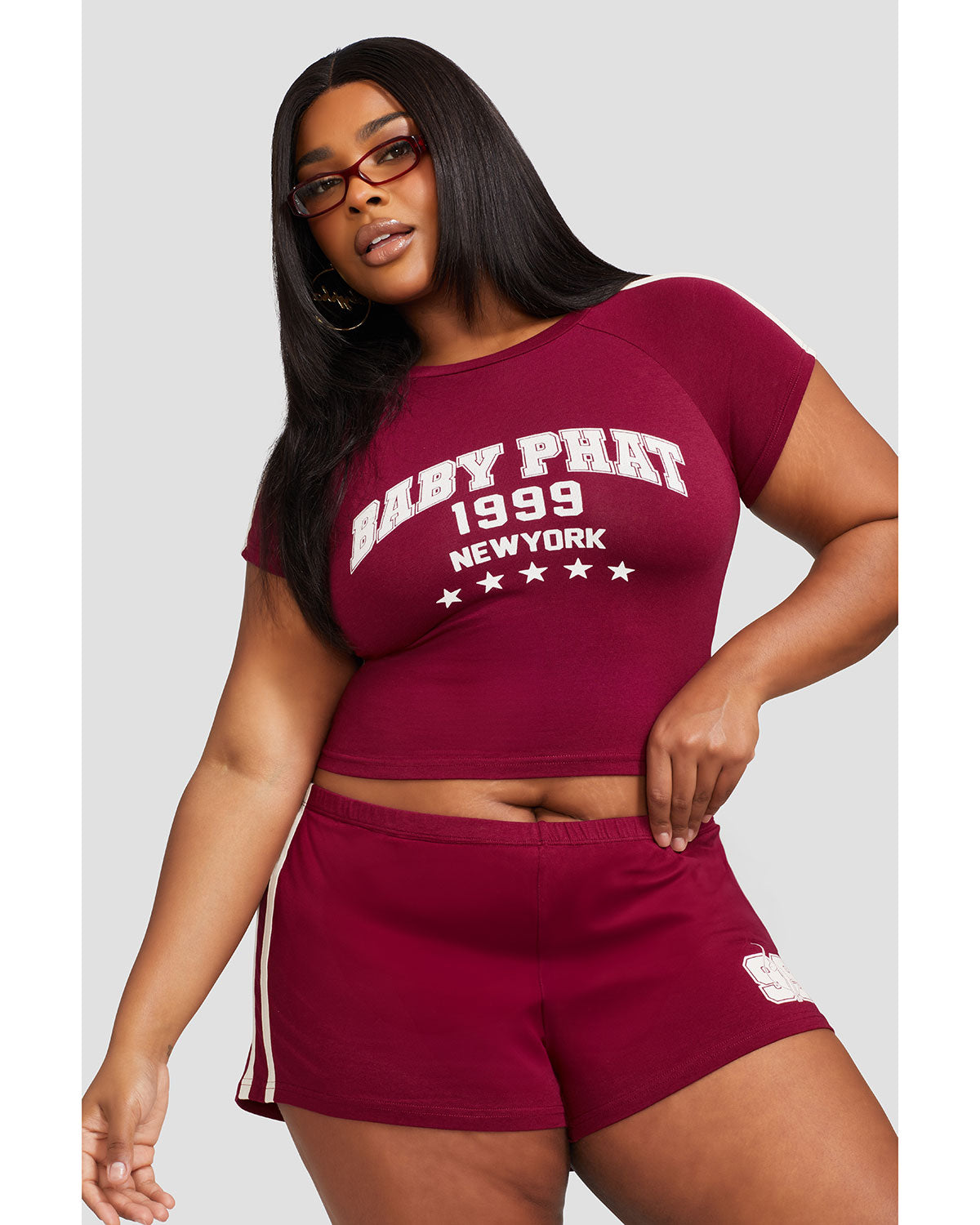 Plus sized model shown wearing burgundy baby tee shirt with cream Baby Phat logo, New York, 1999, and stars on the center front. Shirt features cream stripes on the shoulder. Model is also wearing matching burgundy micro shorts with cream side stripes and a 99 cat logo.