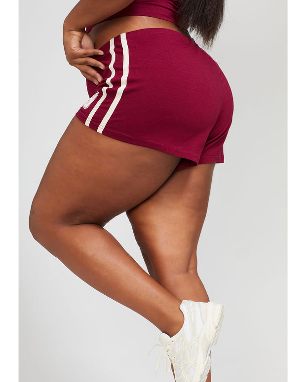 Side view image of plus sized model’s legs shown wearing burgundy micro short with 2 contrasting off white stripes down both sides. Short features off white Baby Phat cat 99 heritage logo on wearer's left thigh. 
