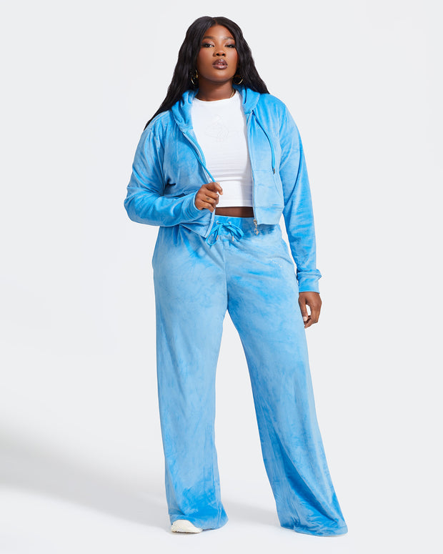 Full length image of plus sized model shown wearing blue velour sweatpants. Pants feature a wide leg, open bottom fit with a rhinestone Baby Phat cat logo on the wearer's left upper thigh. Pants feature pockets and an elastic waistband with a drawstring tie. Model is also wearing a matching blue zippered sweatshirt unzipped to show white baby tee.